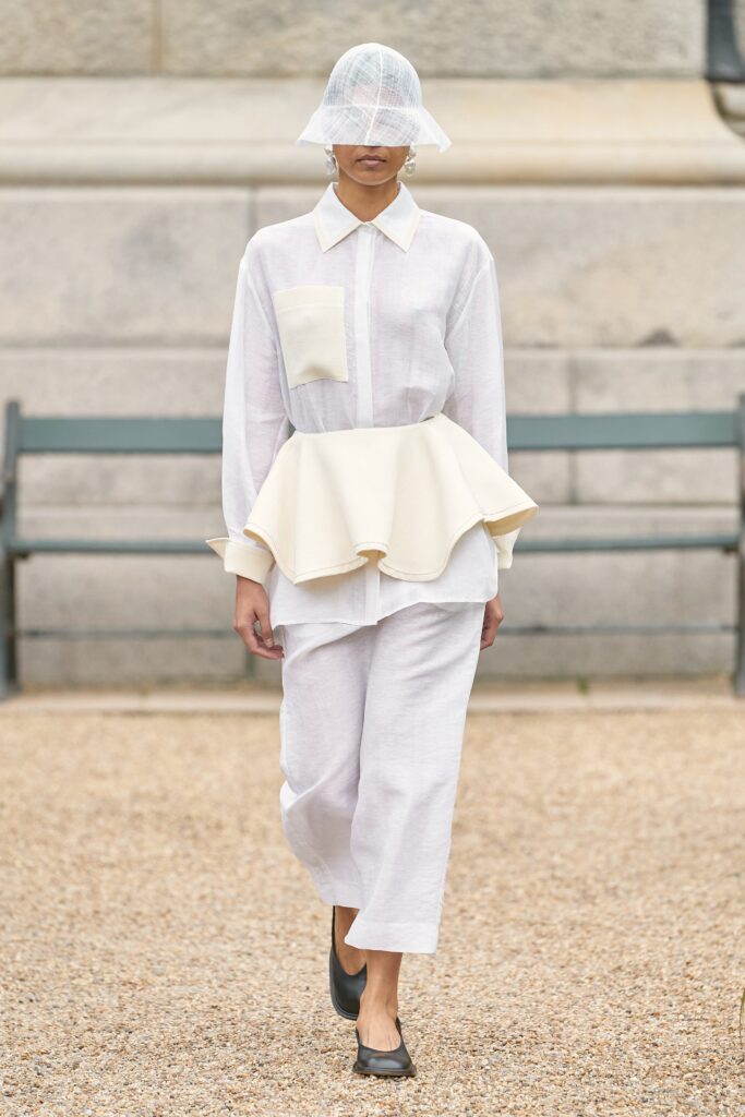 A relaxed white long-sleeve top with a pleated asymmetric design paired with high-waisted beige pants.