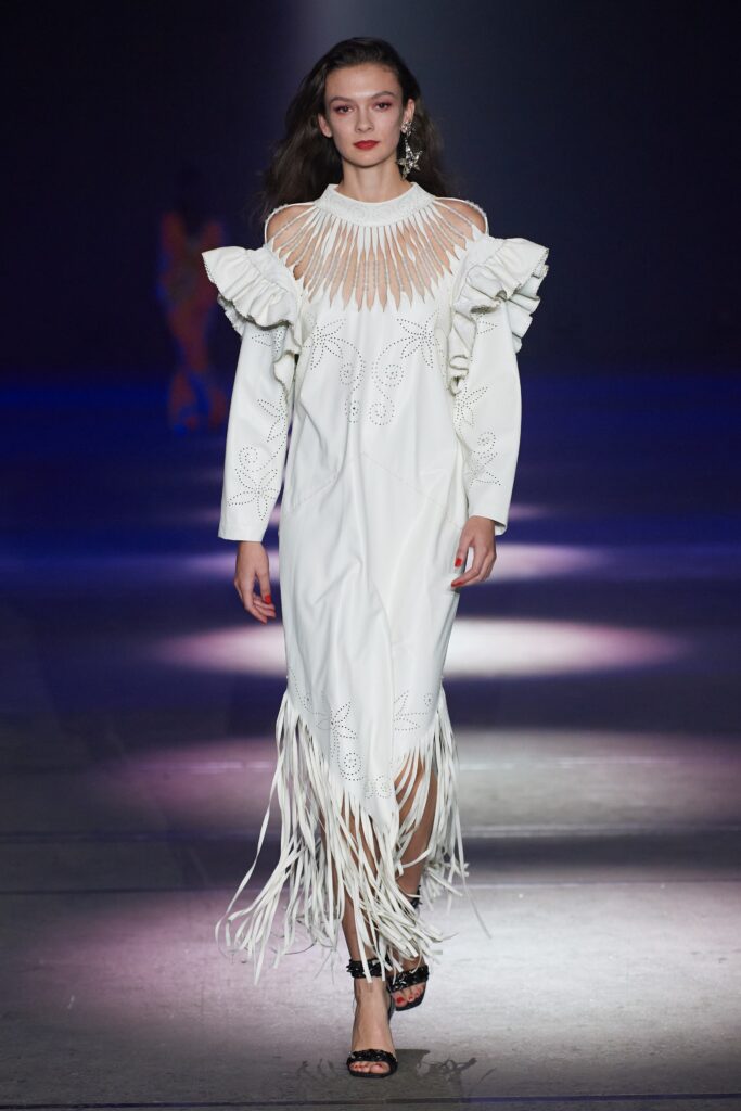 A fitted white dress featuring long sleeves, a high neckline, ruffled details, and fringed accents.