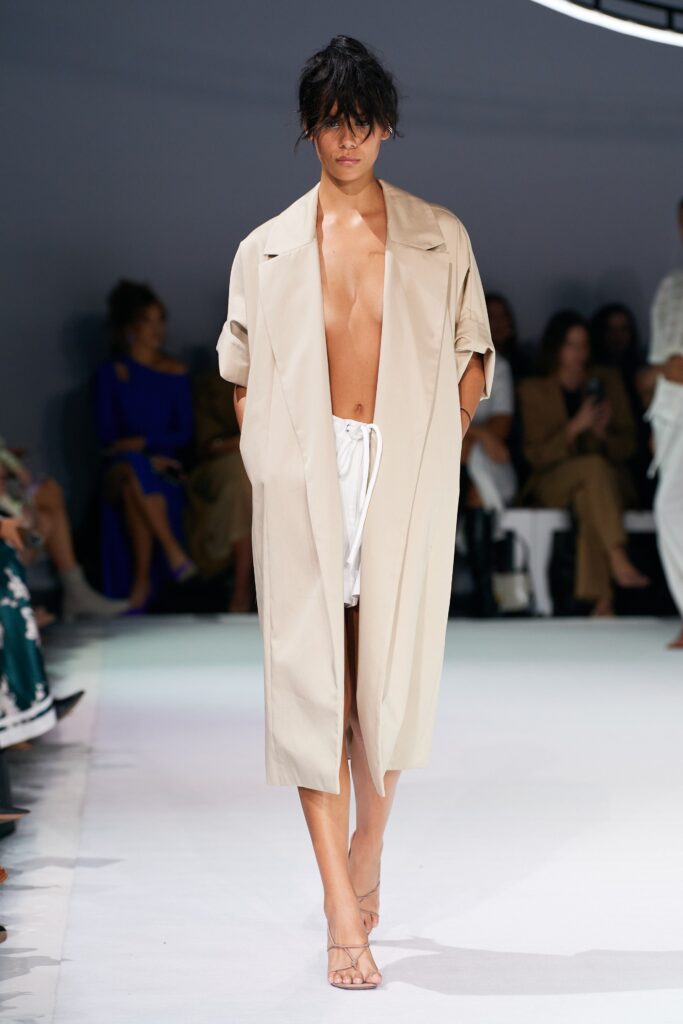 A relaxed beige cotton coat paired with casual shorts, featuring a collared design and buttoned details.