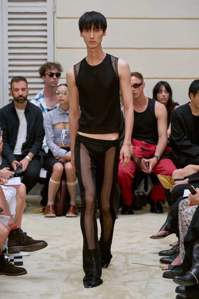 A fitted black sheer sleeveless top with high-neck and asymmetric design paired with sheer pants.