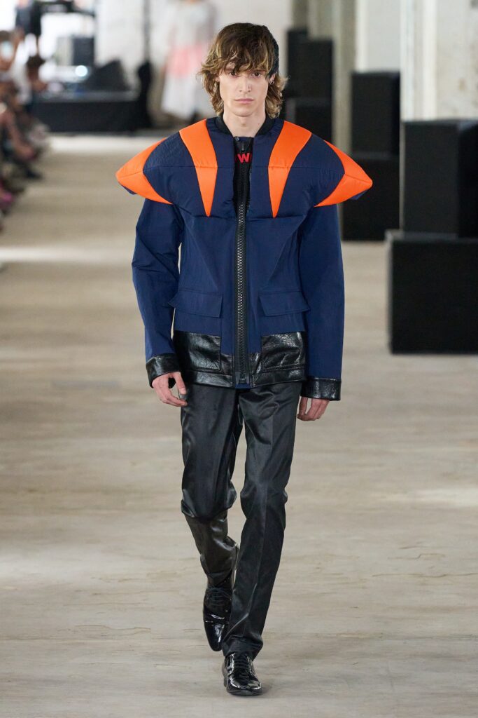 A fitted blue jacket with orange accents, paired with black leather pants and an asymmetric design.