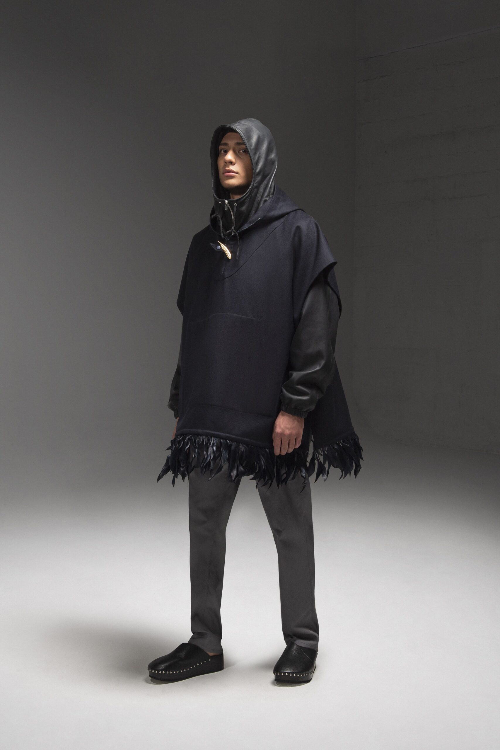 A relaxed black wool hooded poncho with fringed detailing, paired with gray pants.
