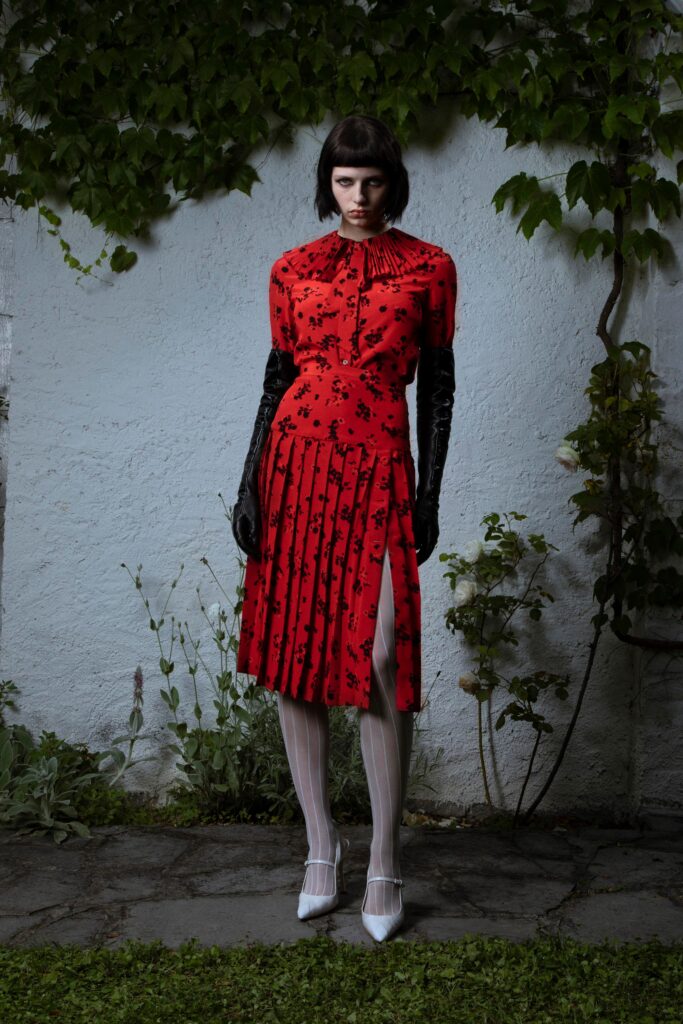 A fitted red silk dress with long sleeves, a V-neckline, pleated design, and a side slit.