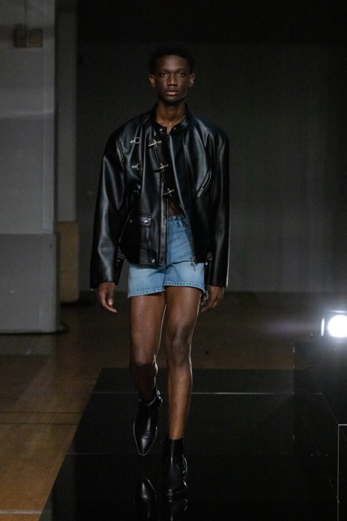 A fitted black leather jacket paired with blue denim shorts, featuring an asymmetric design.