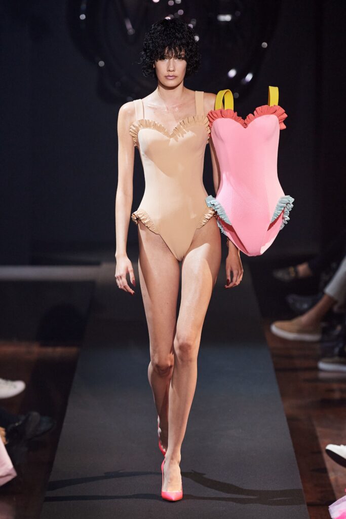 A fitted beige swimsuit with ruffled details and a low-cut design, complemented by a pink accessory.