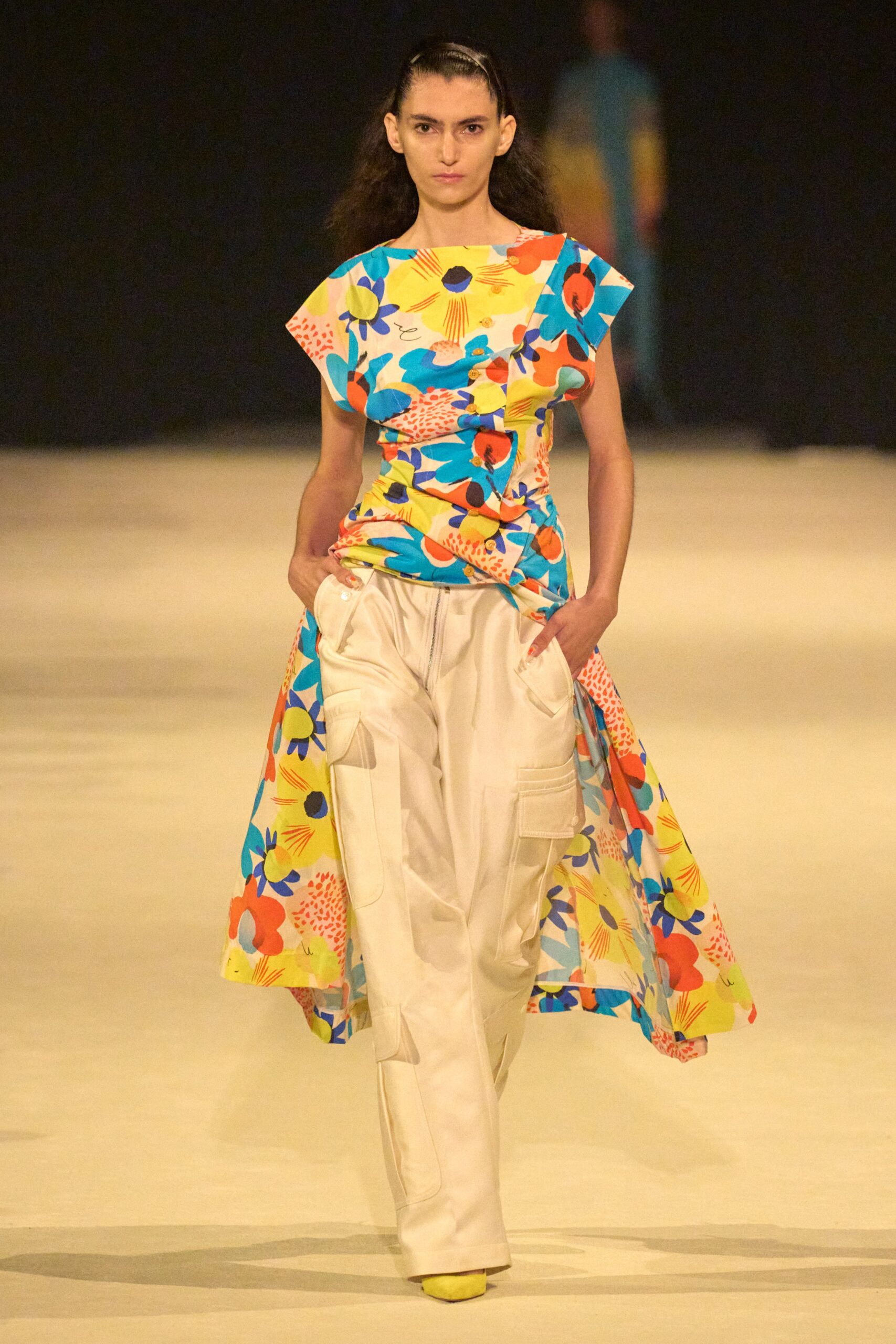 A relaxed multicolor sleeveless dress with an asymmetric design paired with white pants.