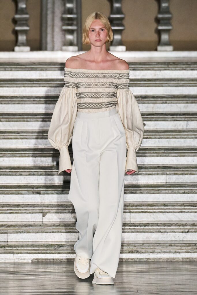 A relaxed white off-shoulder top with long sleeves paired with pleated pants.