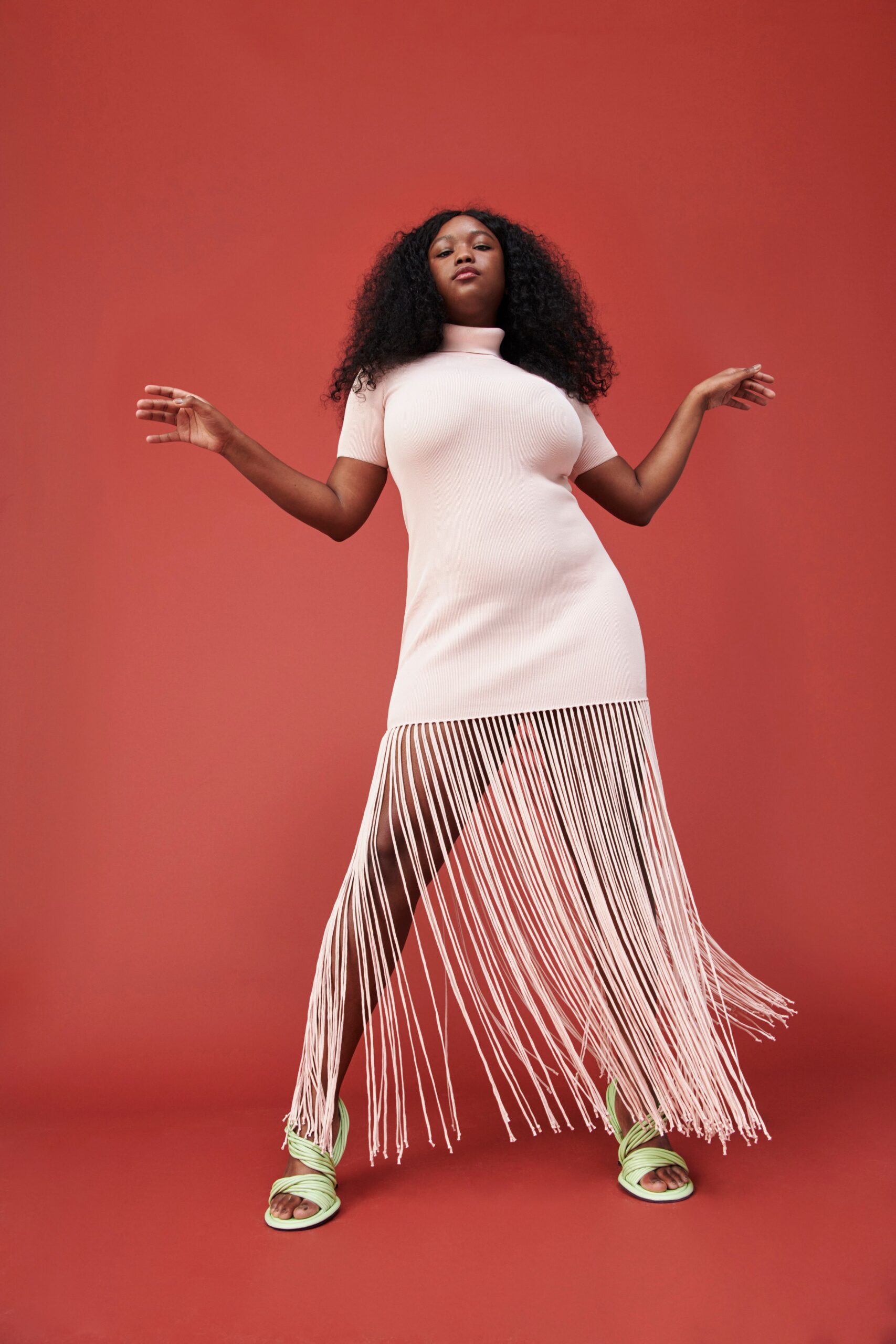 A fitted pink dress with short sleeves, a high neckline, and an asymmetric fringed hem.