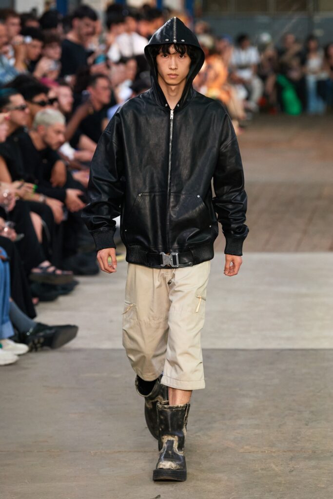A relaxed black leather hooded jacket paired with beige cotton pants featuring a zippered detail.