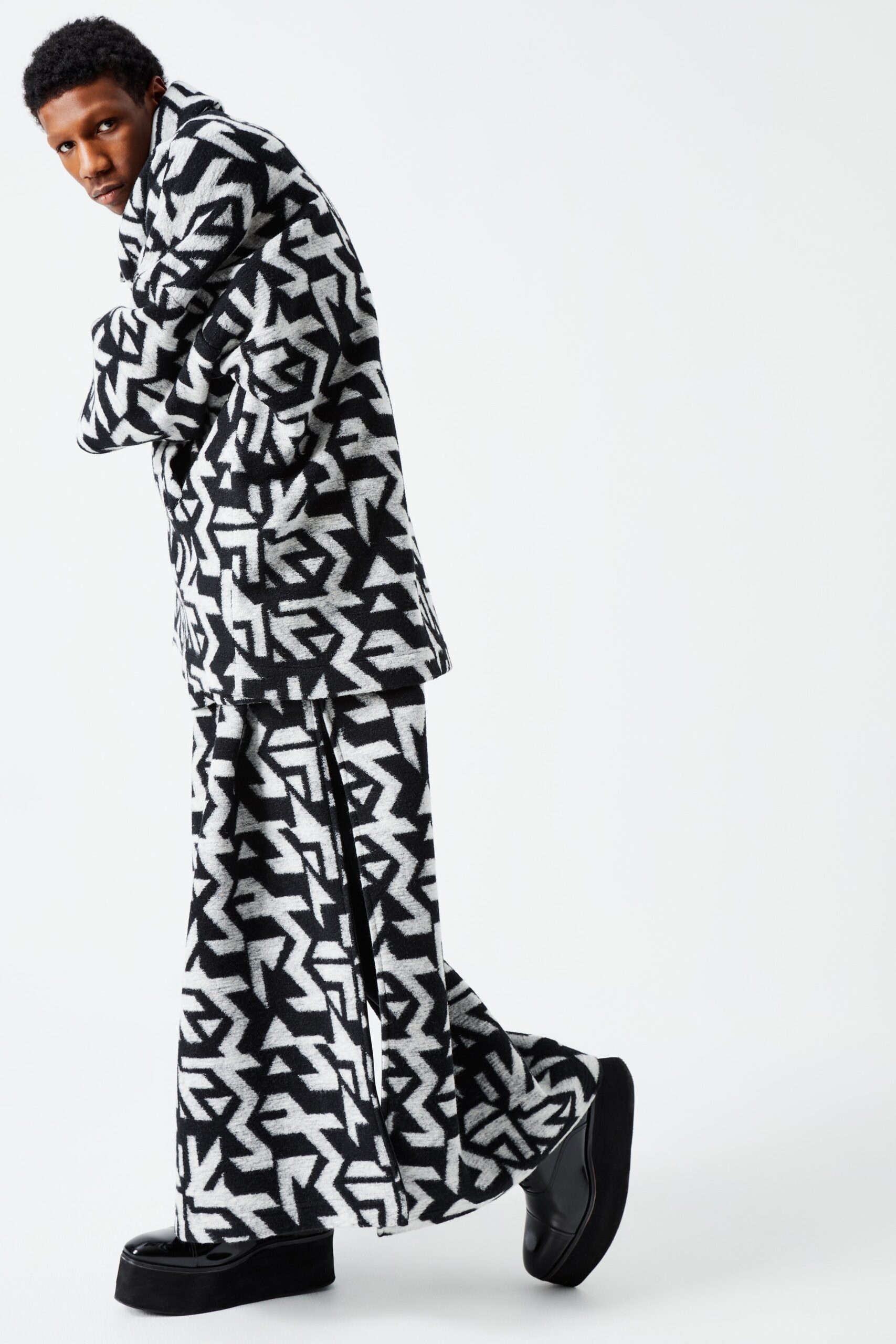 This ensemble features a relaxed-fit jacket and wide-leg pants, both adorned with a striking black and white geometric print. The long sleeves and collared neckline of the jacket add a structured element, while the flowing silhouette of the pants enhances movement and visual interest.