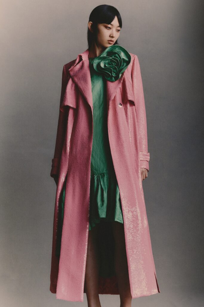A fitted pink silk dress with an asymmetric design, layered under a long coat.