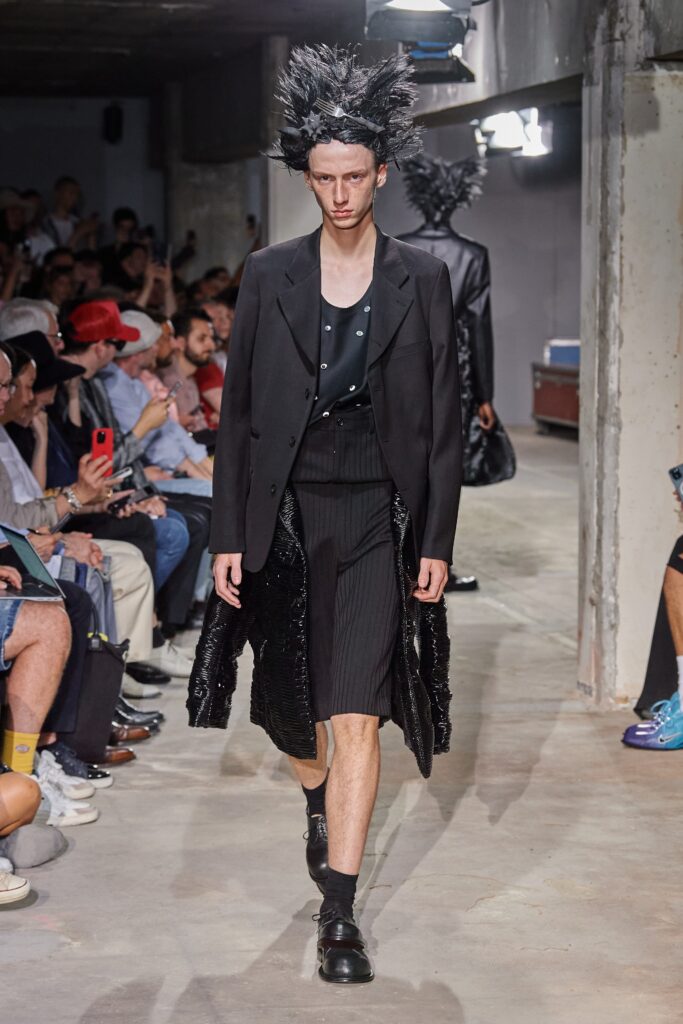 A structured black wool suit featuring long sleeves, a collared design, and an asymmetric detail.