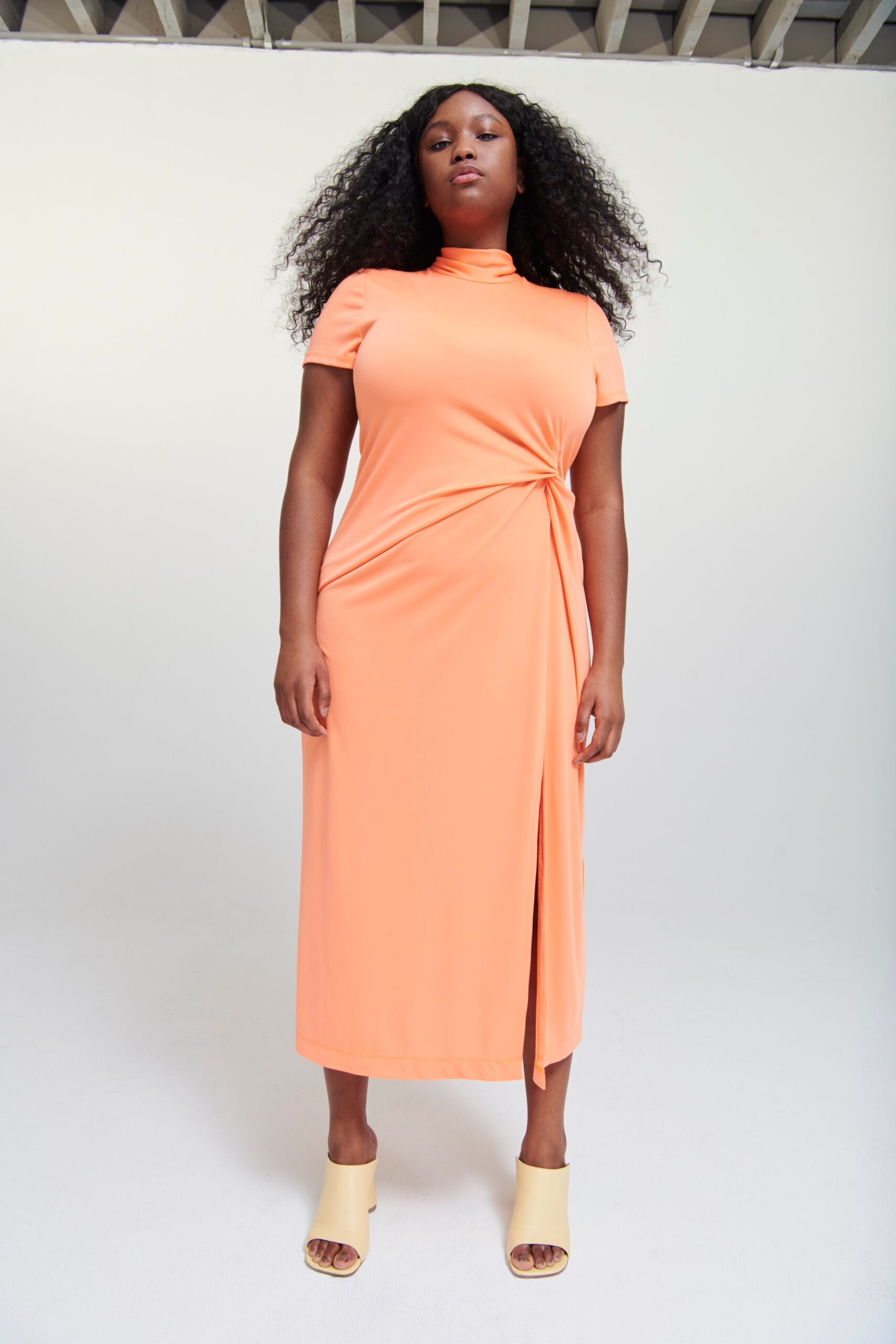 A fitted orange jersey dress with short sleeves, a high neckline, and an asymmetric slit detail.