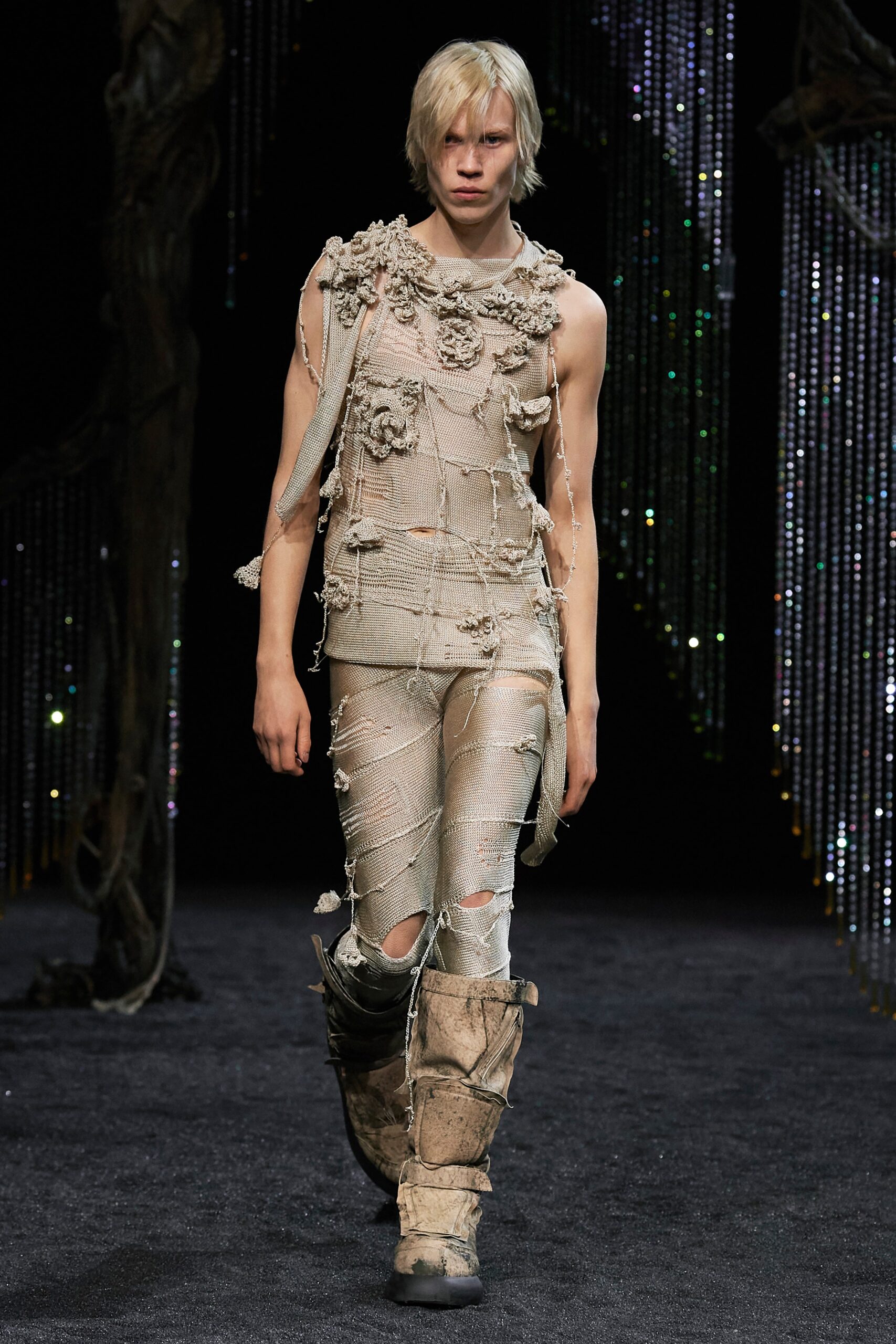 A fitted beige sleeveless top with a V-neckline and fringed layered details paired with fitted pants.