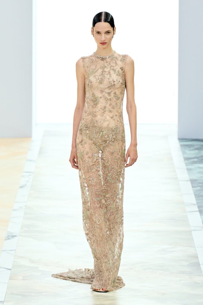 A fitted beige lace gown with a sleeveless design and high-neck construction, featuring intricate embroidery.