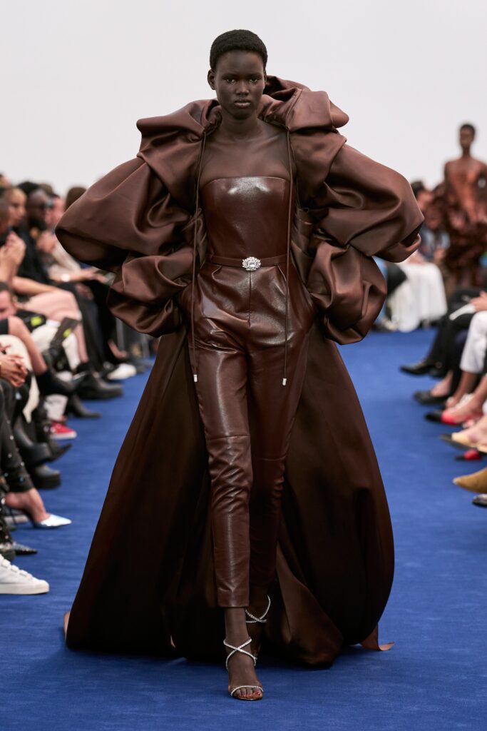 A fitted brown leather jumpsuit with a high-neck and layered cape detail.