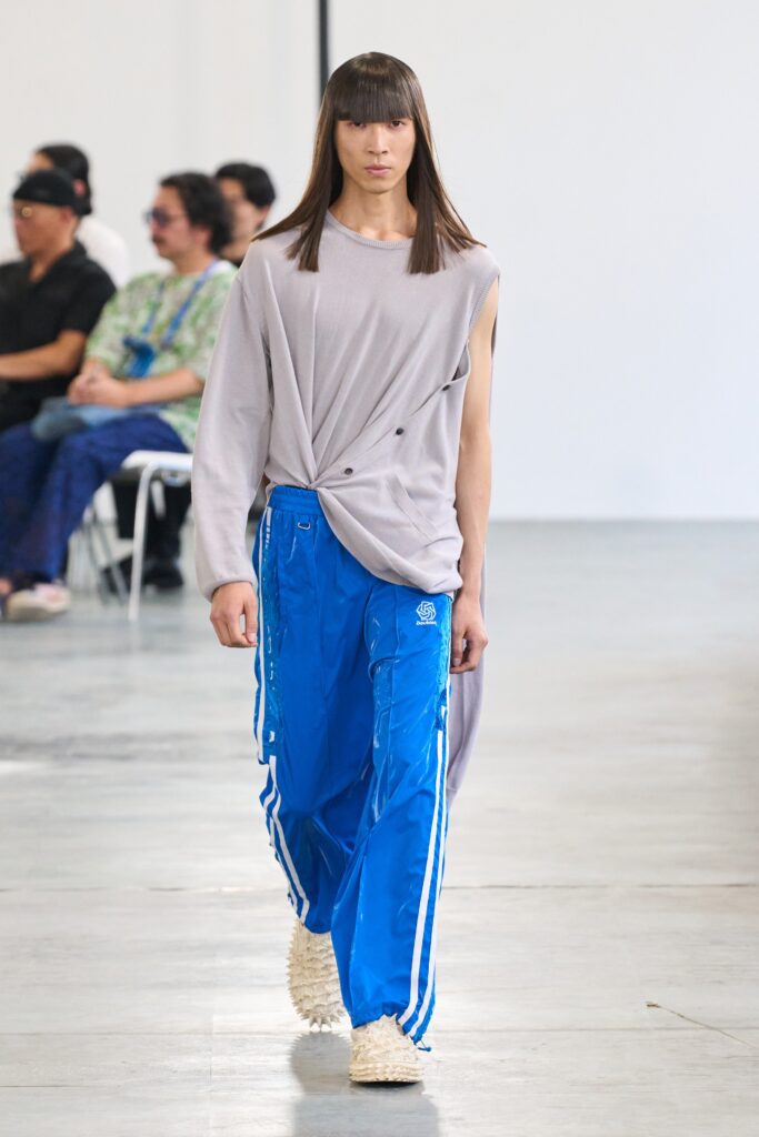A relaxed gray asymmetric long-sleeve top paired with blue pants featuring a zippered detail.