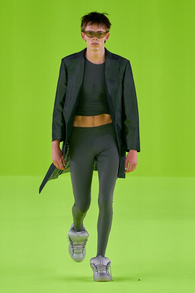 A fitted black jacket with long sleeves and asymmetric design paired with gray pants.