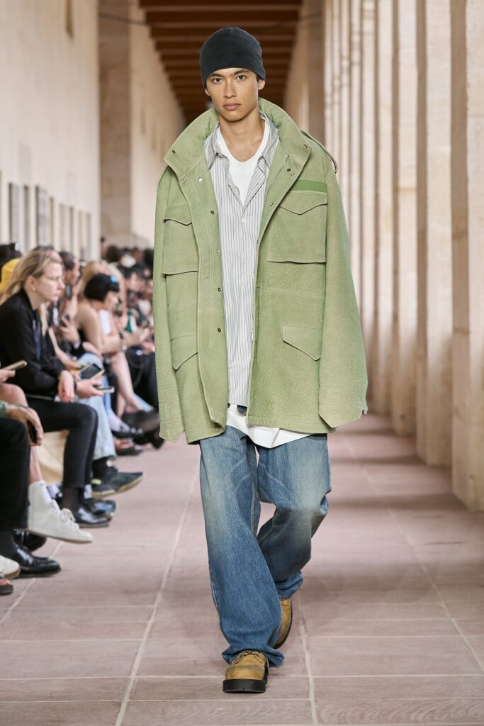 A relaxed green jacket layered over a collared white shirt and blue denim pants.