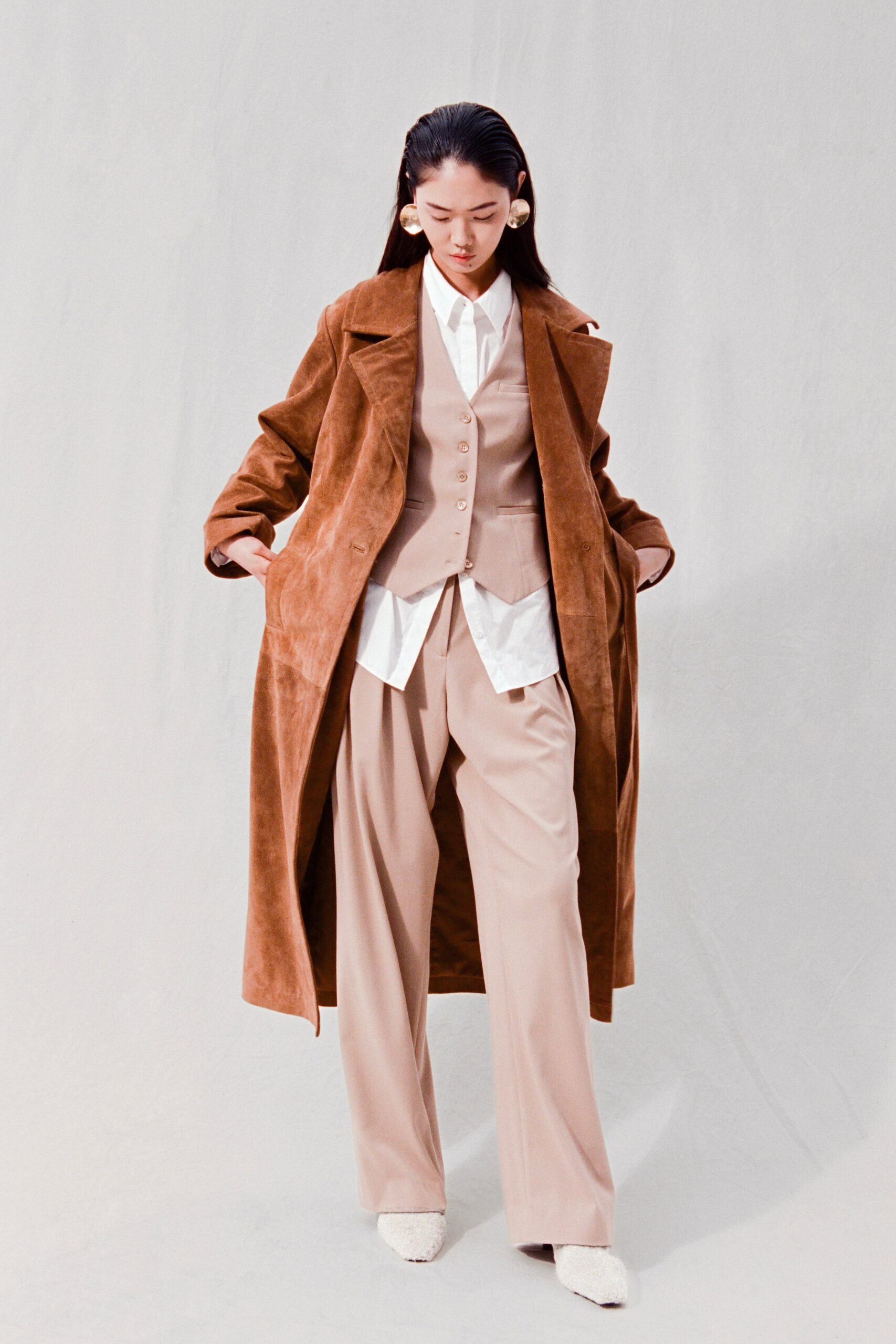 A relaxed brown suede coat layered over a beige vest and wide-leg trousers.