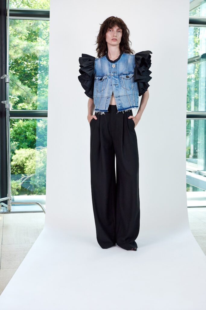 A relaxed blue denim sleeveless jacket with ruffled details paired with wide-leg black pants.