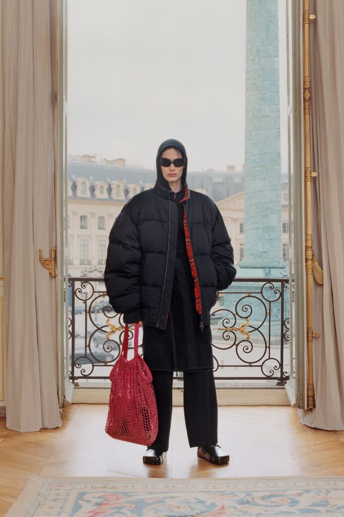 An oversized black coat with a hood, paired with black pants and a red accent.