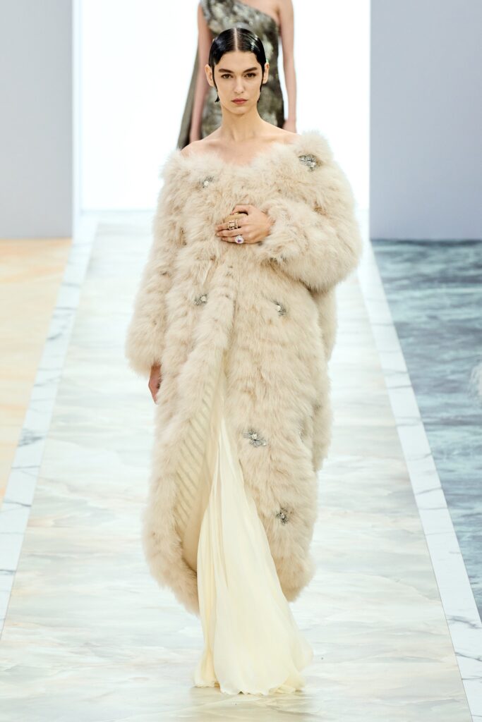 A fitted cream fur coat worn over a sleeveless silk gown with a high neckline.