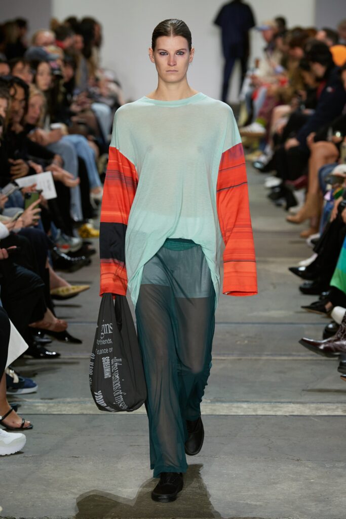 A relaxed blue sheer long-sleeve top layered over green pants, featuring bold red accents.
