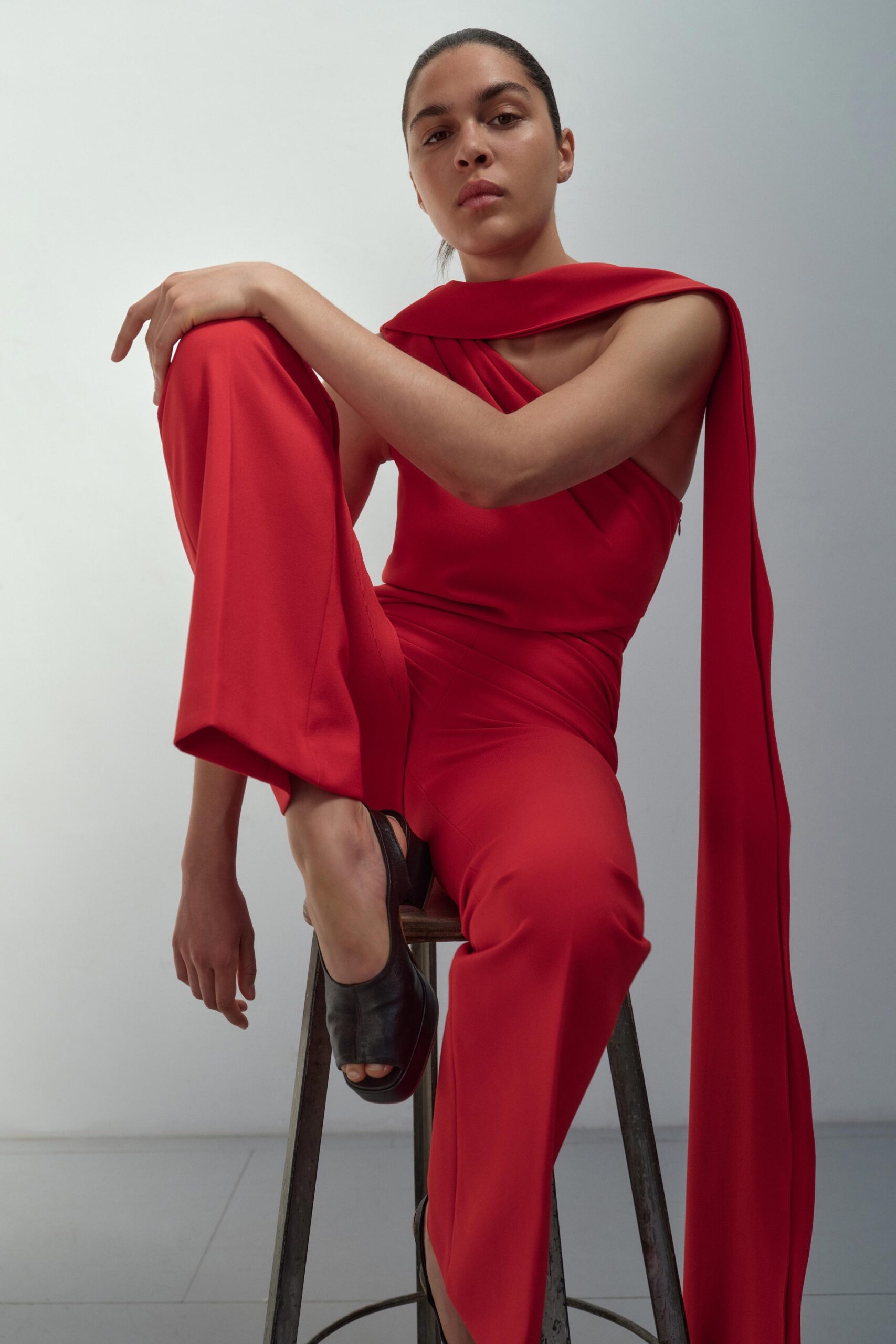 A fitted red sleeveless jumpsuit with a high-neck and asymmetric design detail.