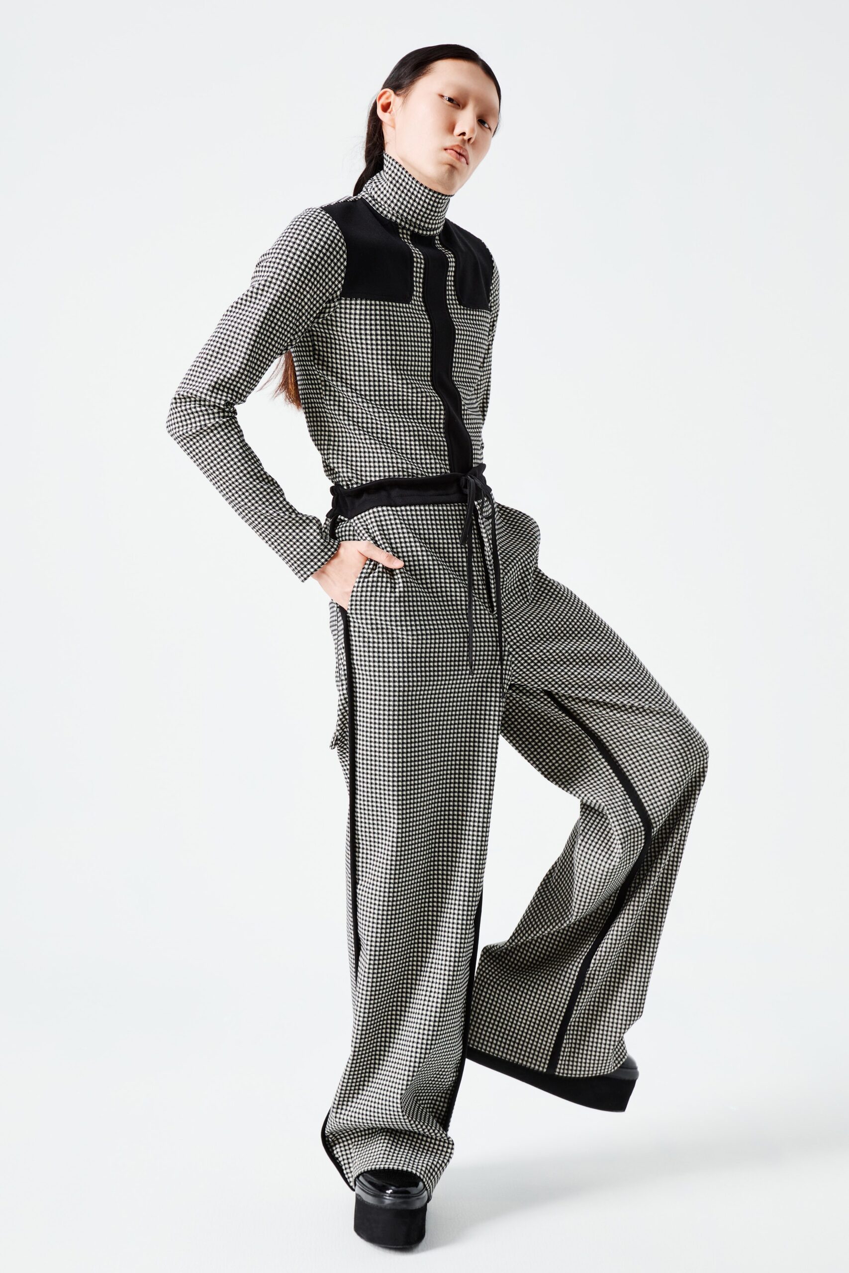 This fitted jumpsuit showcases a striking black and white checked pattern, accentuated by a structured collar and a zippered front. The long sleeves and tailored silhouette create a modern, streamlined look that emphasizes the bold graphic elements.