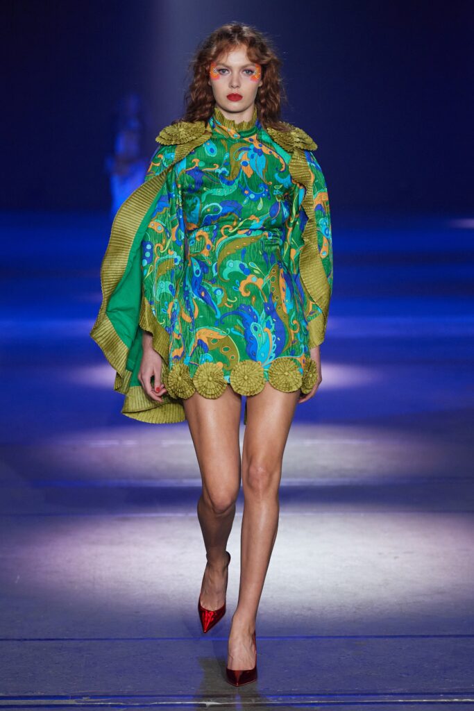 A fitted green silk dress with a high-neck and flared silhouette, complemented by a vibrant cape.