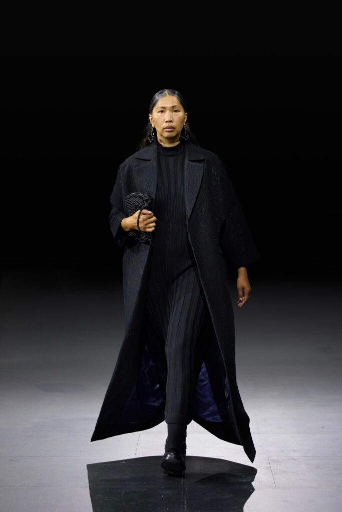 An oversized black wool coat layered over a fitted black jumpsuit with long sleeves.