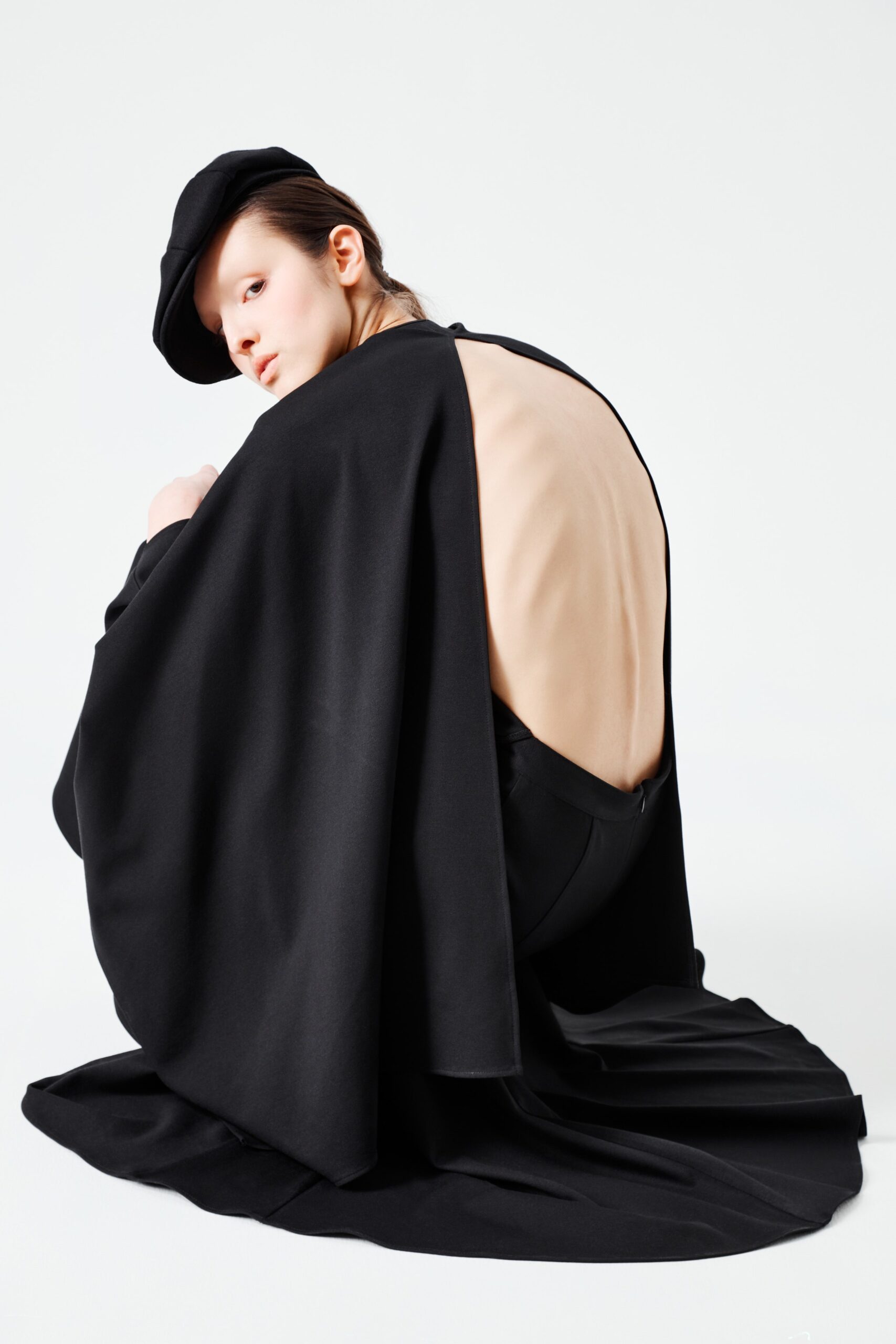 This striking black dress features a fitted silhouette that accentuates the form, complemented by a dramatic cape that flows elegantly behind. The backless design adds an unexpected twist, creating a captivating interplay of structure and movement.