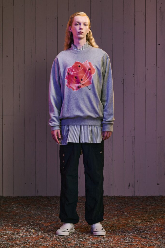 A relaxed gray sweater with a printed design layered over a collared shirt, paired with black pants.