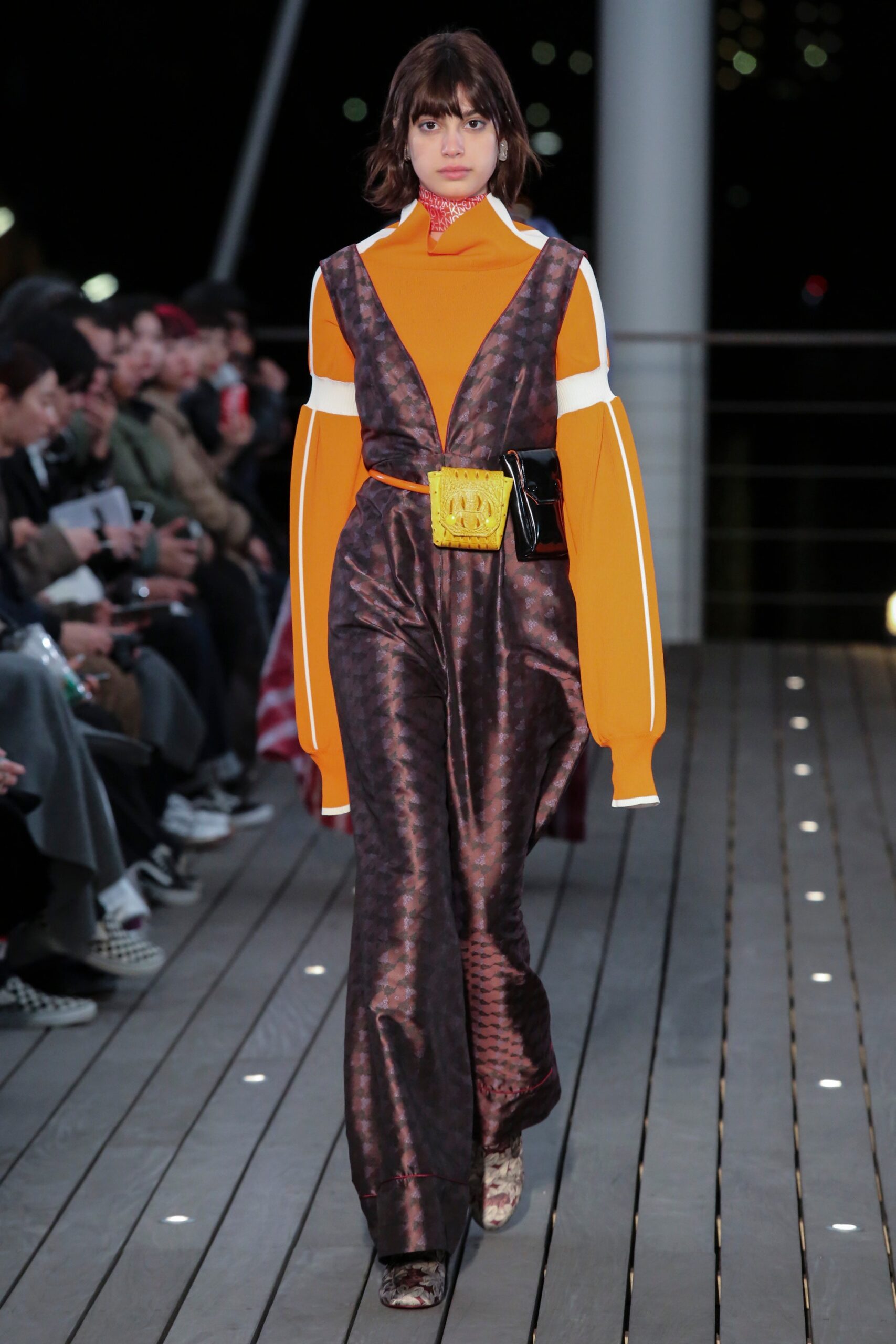 A fitted orange and brown jumpsuit with a V-neckline and long sleeves featuring an asymmetric design.