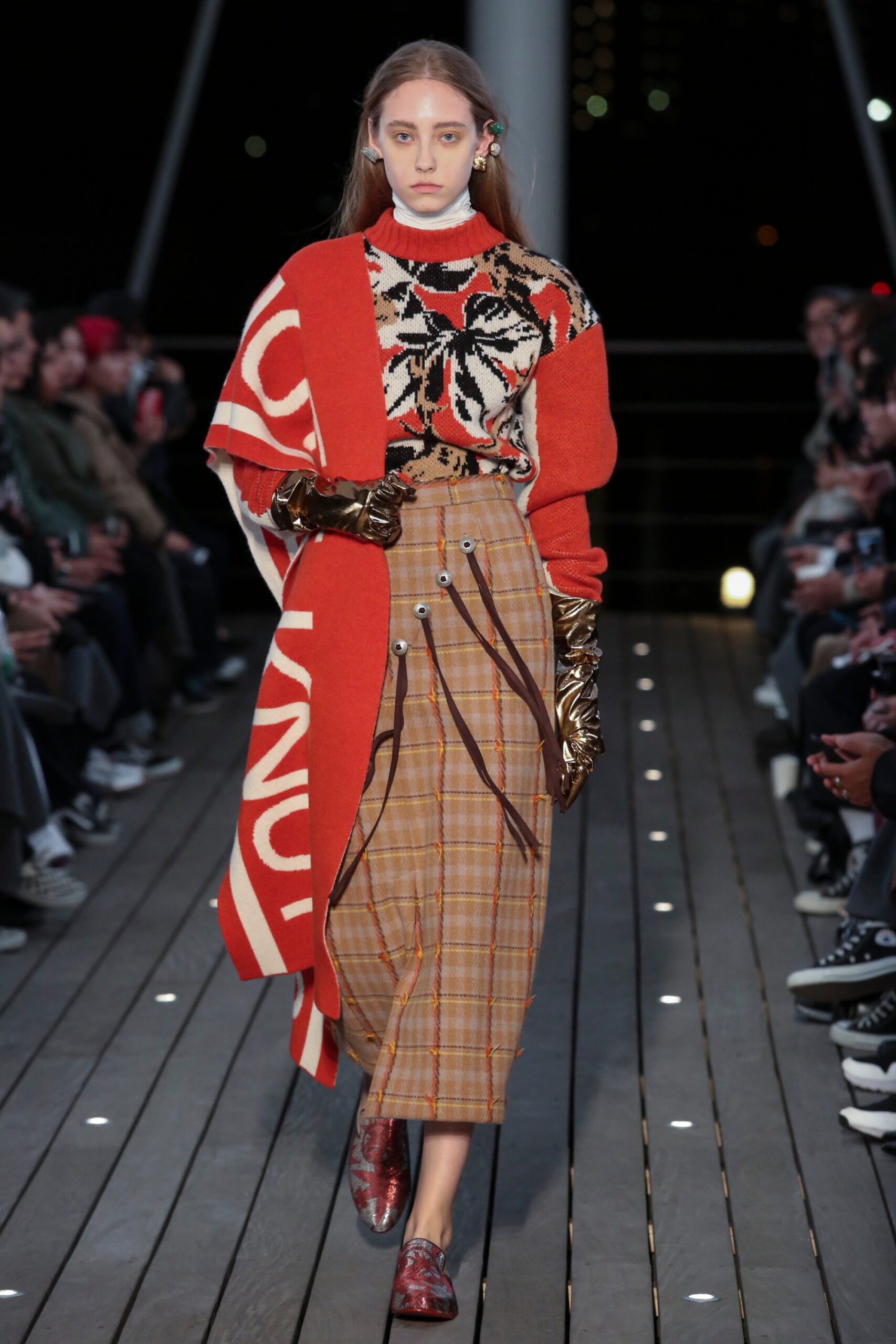 A relaxed red printed sweater paired with a beige pleated skirt, featuring a high neckline and long sleeves.