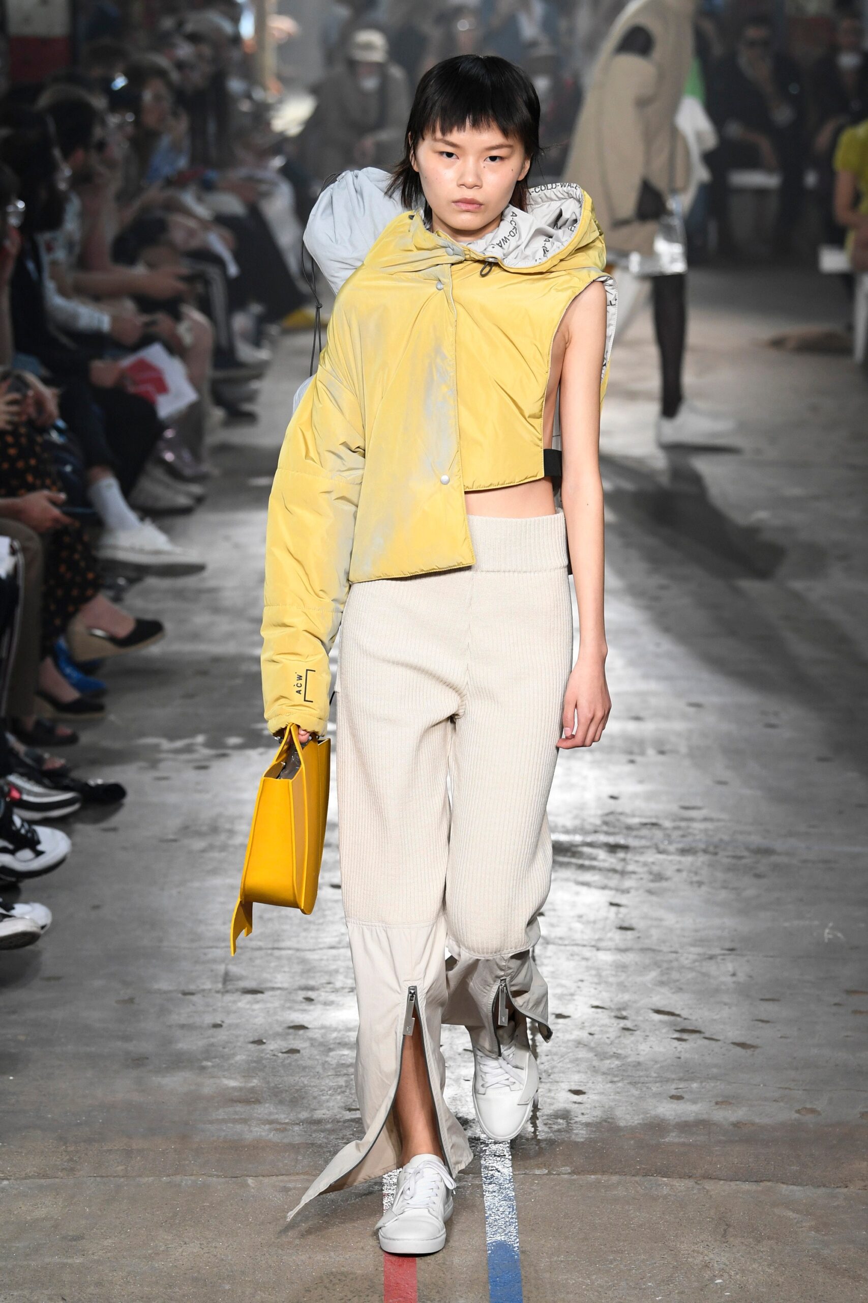 This ensemble features a relaxed yellow jacket with an asymmetric design, showcasing a unique silhouette that draws the eye. Paired with white pants that incorporate zippered details, the look balances casual comfort with contemporary flair, creating an intriguing visual dynamic.