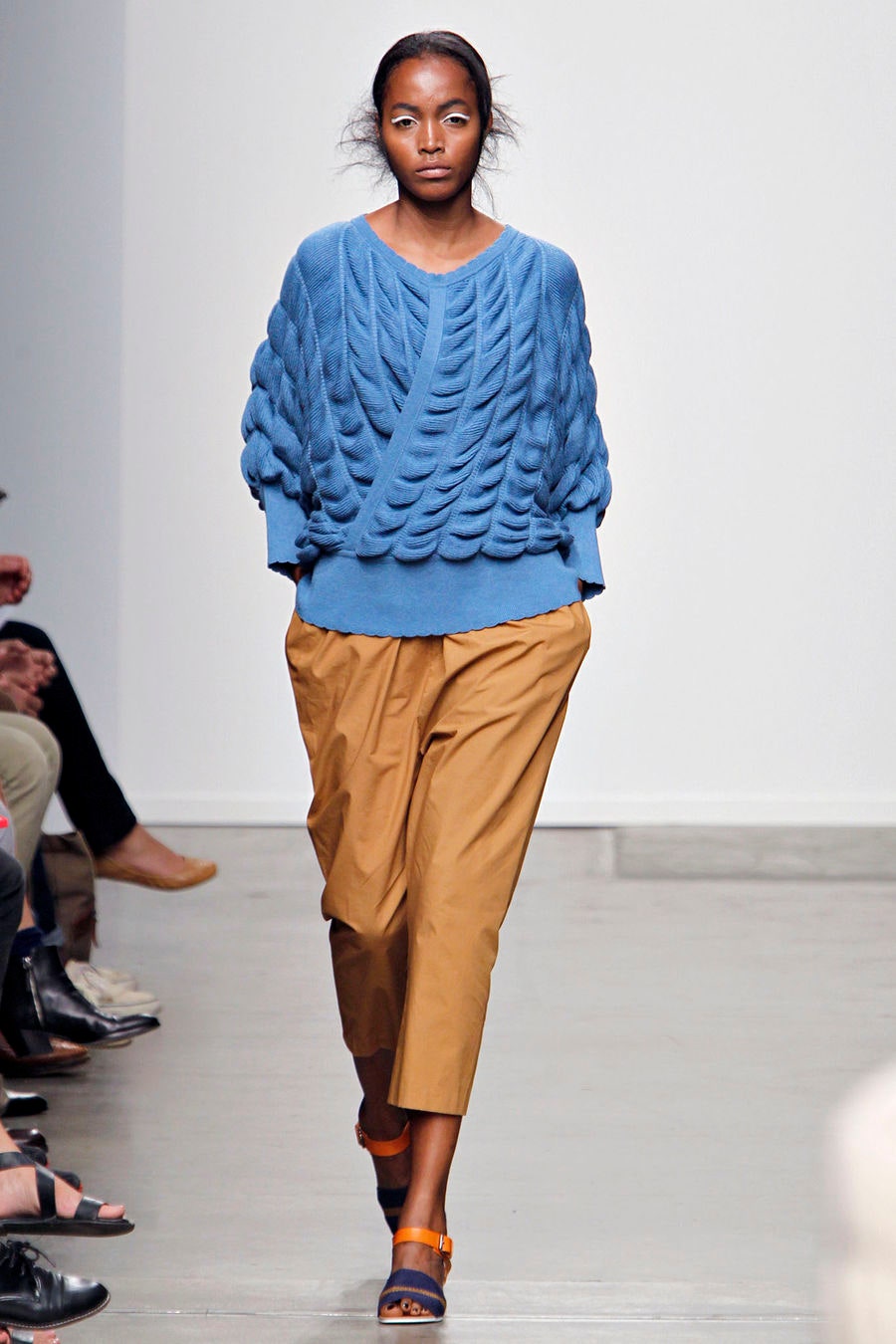 A relaxed blue cotton sweater with a V-neck and pleated design paired with brown pants.