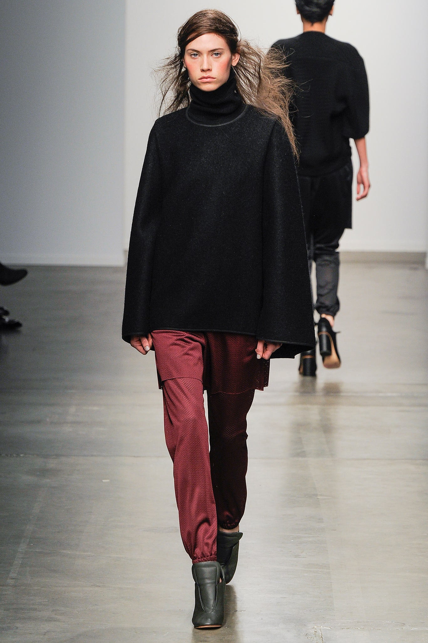 A relaxed black wool cape with a turtleneck and asymmetric design paired with red pants.