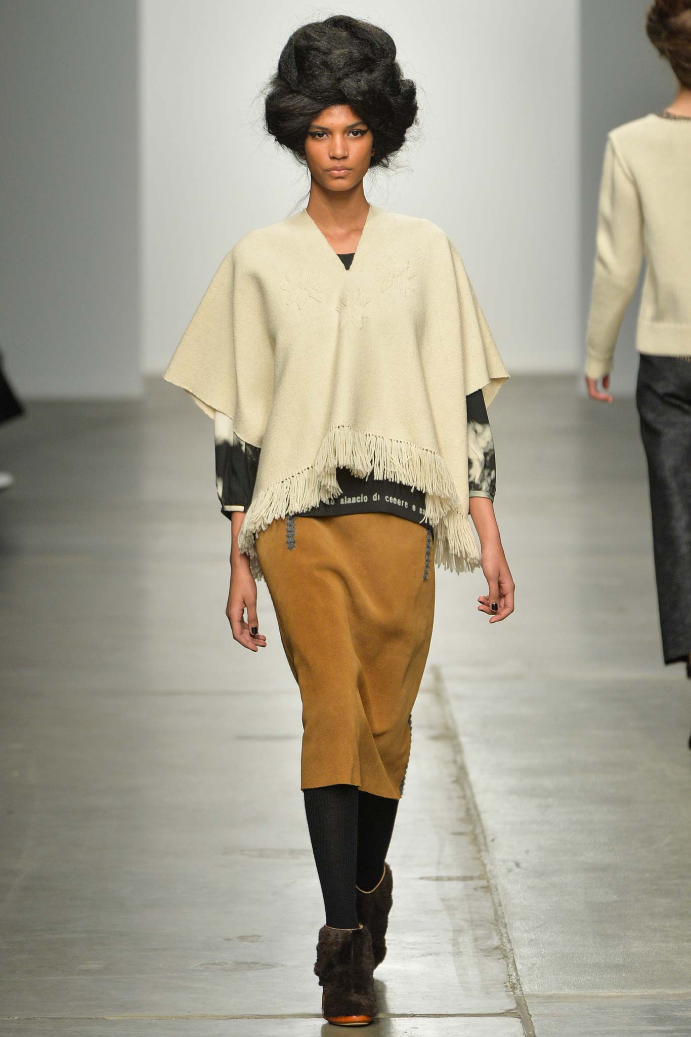 A relaxed beige wool poncho with fringed details paired with a high-waisted brown skirt.