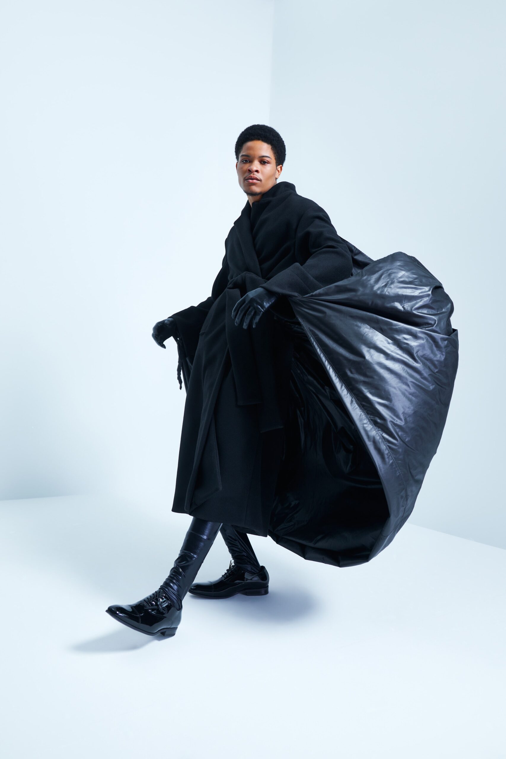 An oversized coat in black leather, featuring a dramatic asymmetric silhouette that flows with movement. The long sleeves and collared design add a structured yet fluid quality, creating a striking visual impact.