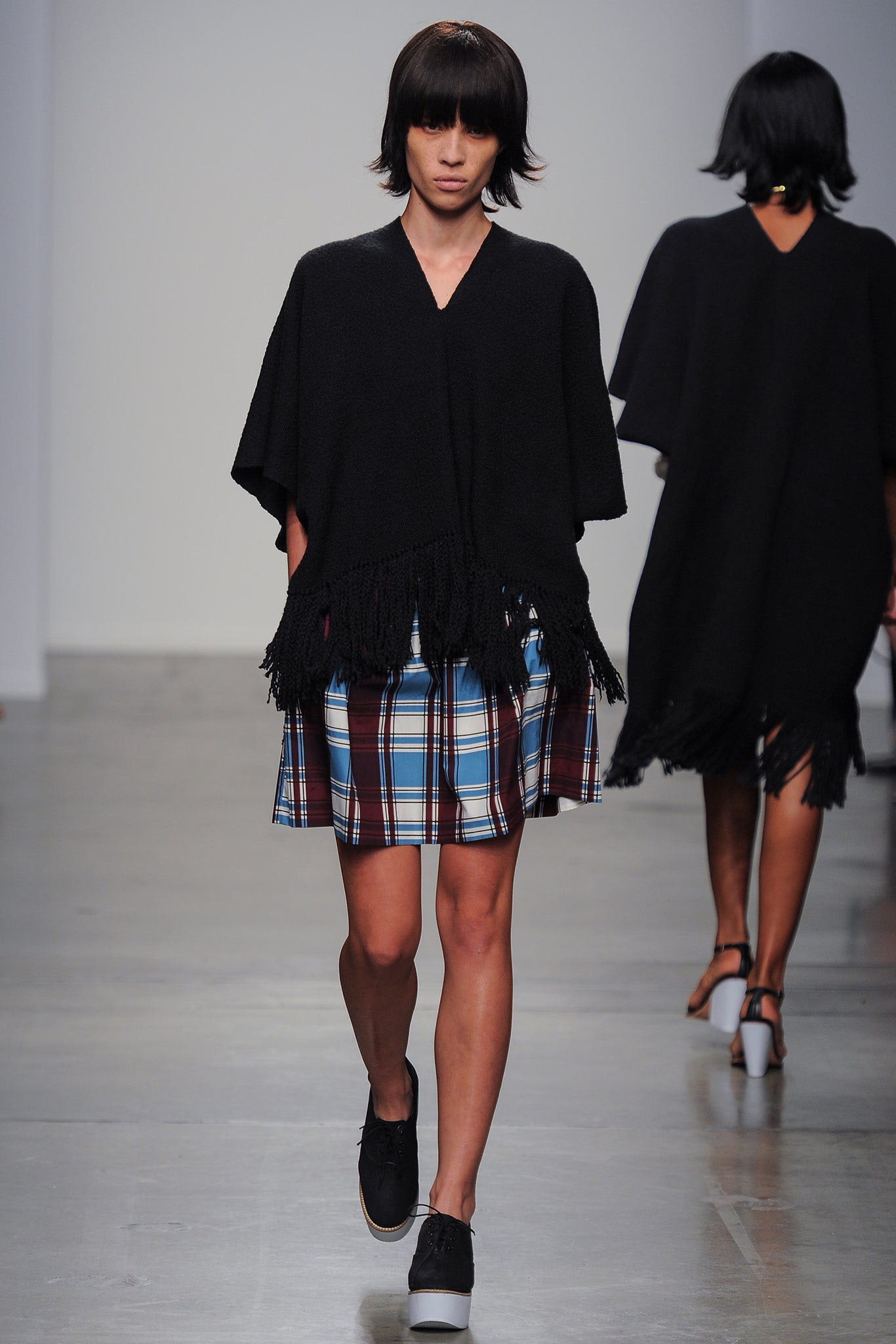 A relaxed black poncho with fringed details paired with a plaid skirt.