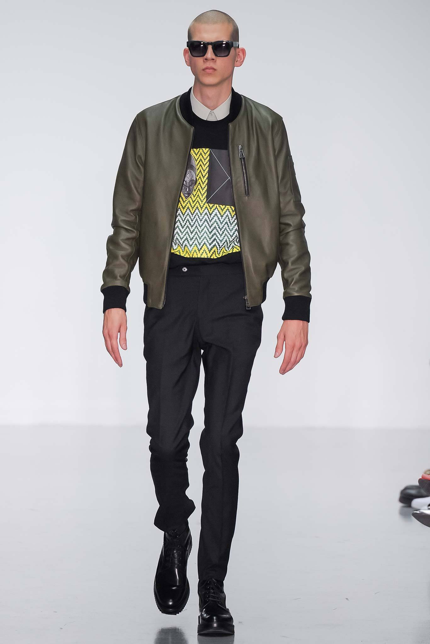 This fitted green leather jacket features a striking zippered front and long sleeves, layered over a vibrant printed sweater that adds a dynamic visual element. The combination of tailored pants completes the look, creating a modern silhouette that balances structure and style.