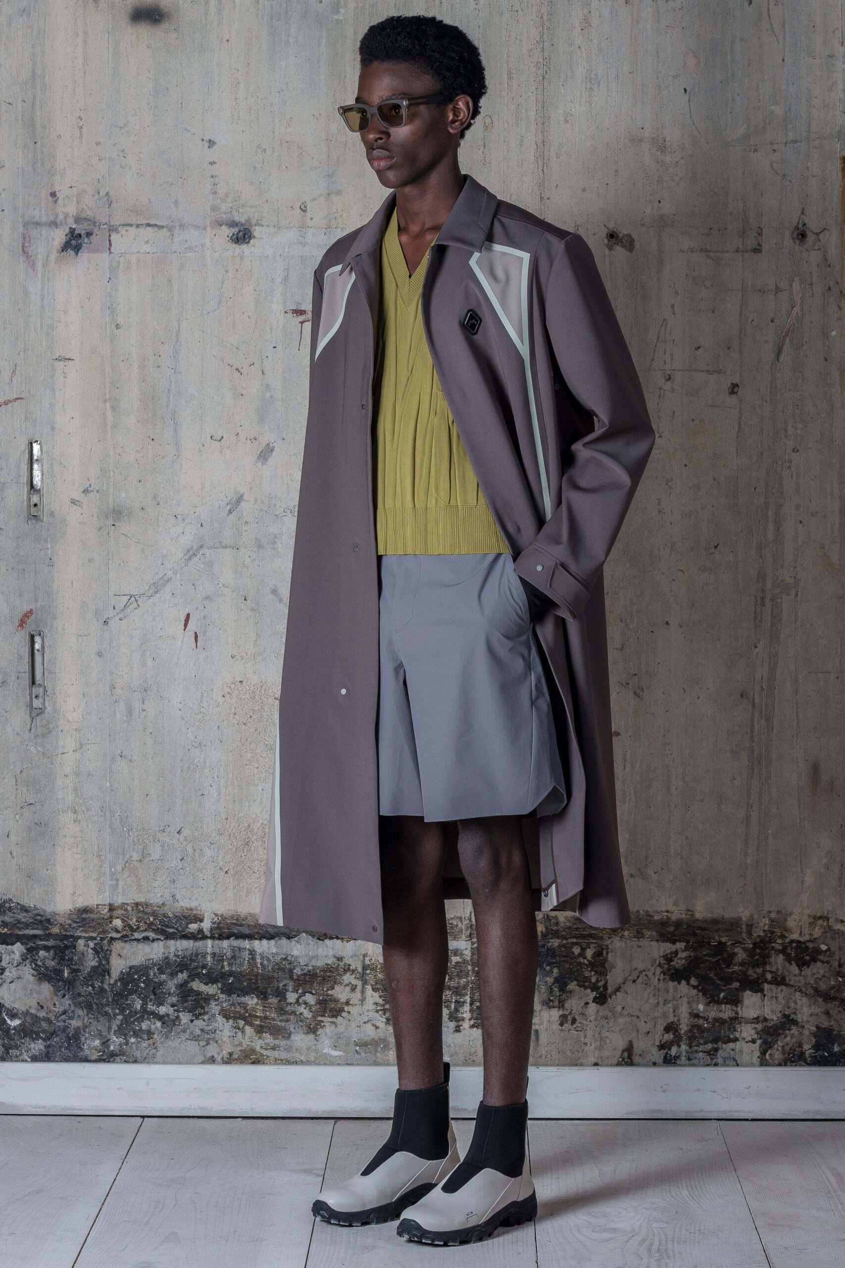 A relaxed long-sleeve coat in gray is layered over a vibrant green v-neck top, creating a striking contrast. The ensemble is complemented by tailored shorts, enhancing the overall silhouette with a modern edge.