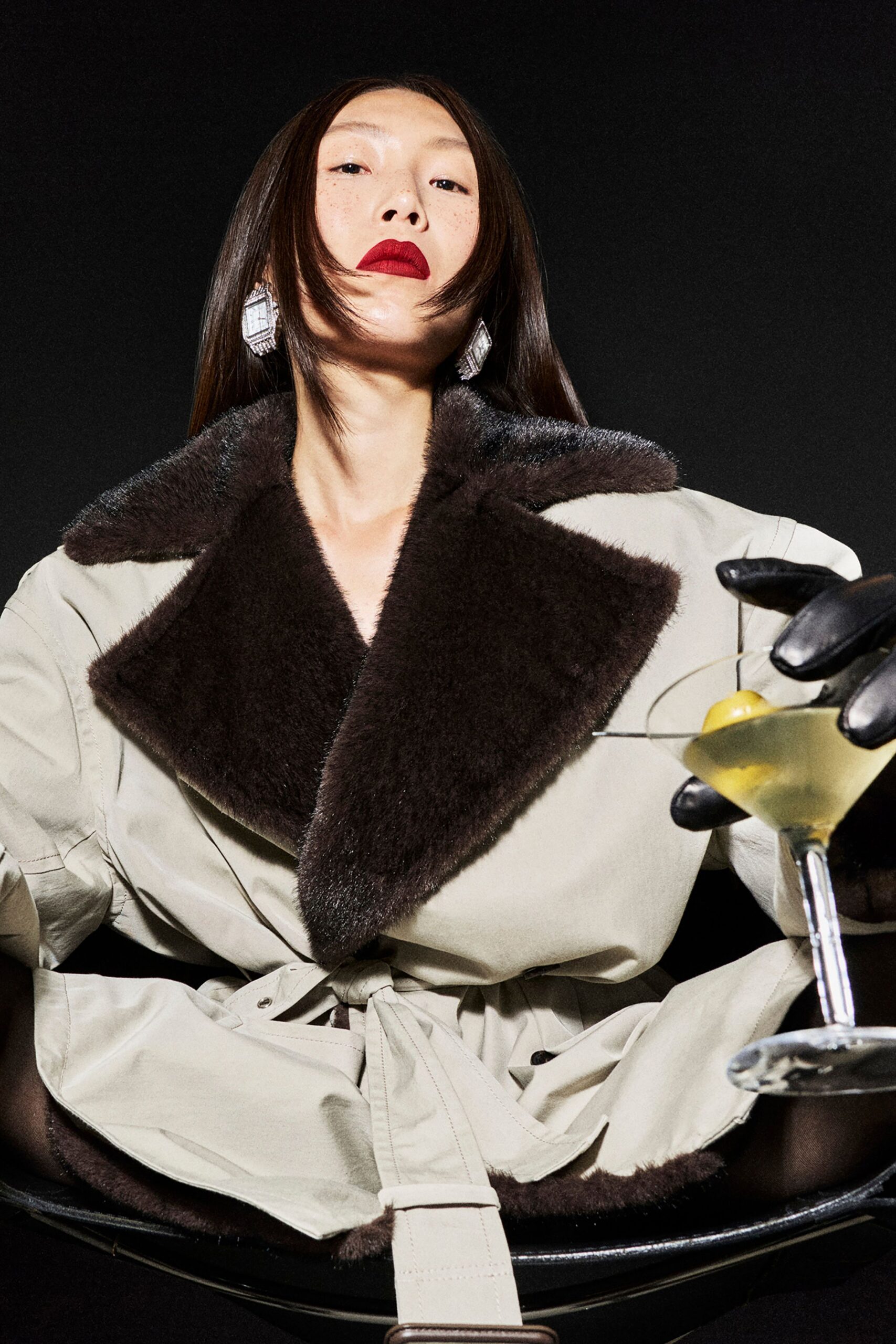 Bone leather trench with an oversized chocolate shearling shawl collar, tied with a self-belt whose tip hangs past the lap. Black leather gloves, faceted crystal drop earrings. Holding a martini that should have stayed at the piano bar.