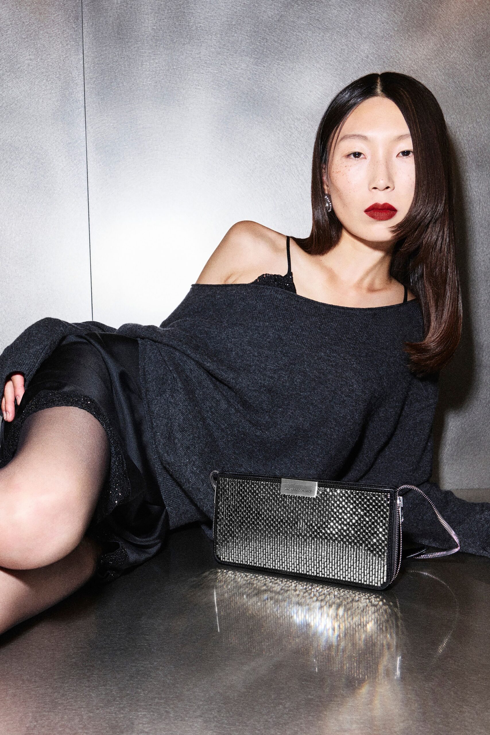 Slouched charcoal knit dress slipping off one shoulder, a black beaded camisole strap catching the light. Sheer tights with a glittered hem. Metal-mesh zip pouch in hand. Waiting out last call in a hotel bar gone quiet.