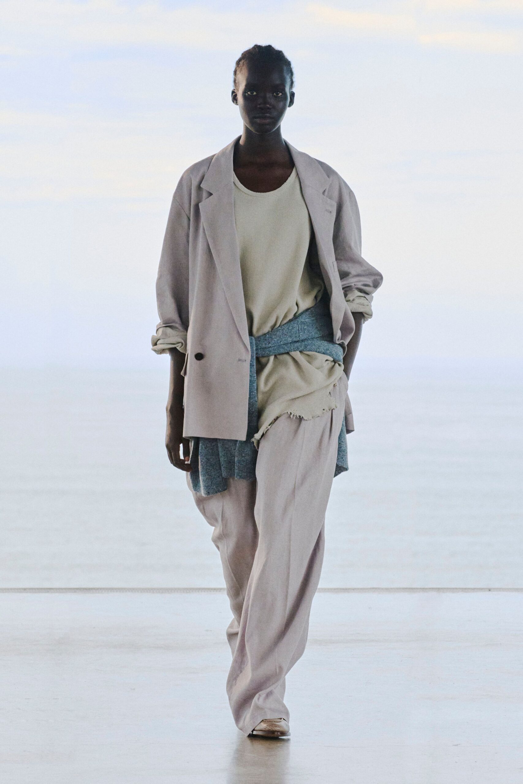 Powder-beige blazer with pushed-up sleeves, over a cream ribbed tank and matching top with a frayed raw hem. Heathered blue knit tied low at the hips, pale loose trousers. Early morning in a near-empty departures hall.