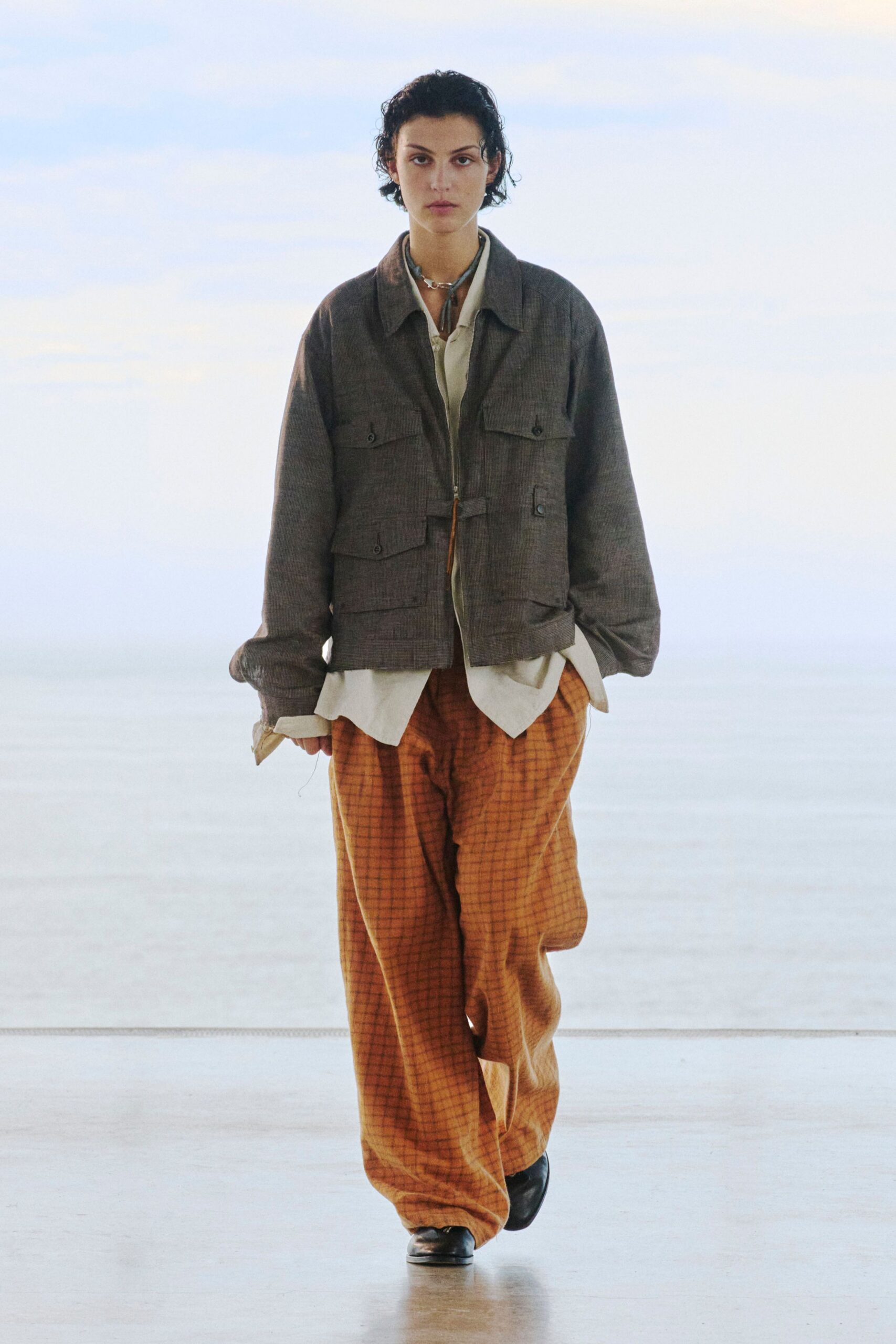 Charcoal zip-front field jacket in fine crosshatch wool, flap pockets set low over an ivory shirt with turned-back cuffs. Rust windowpane trousers break loose over black square-toe shoes. Early train, paper cup gone cold.