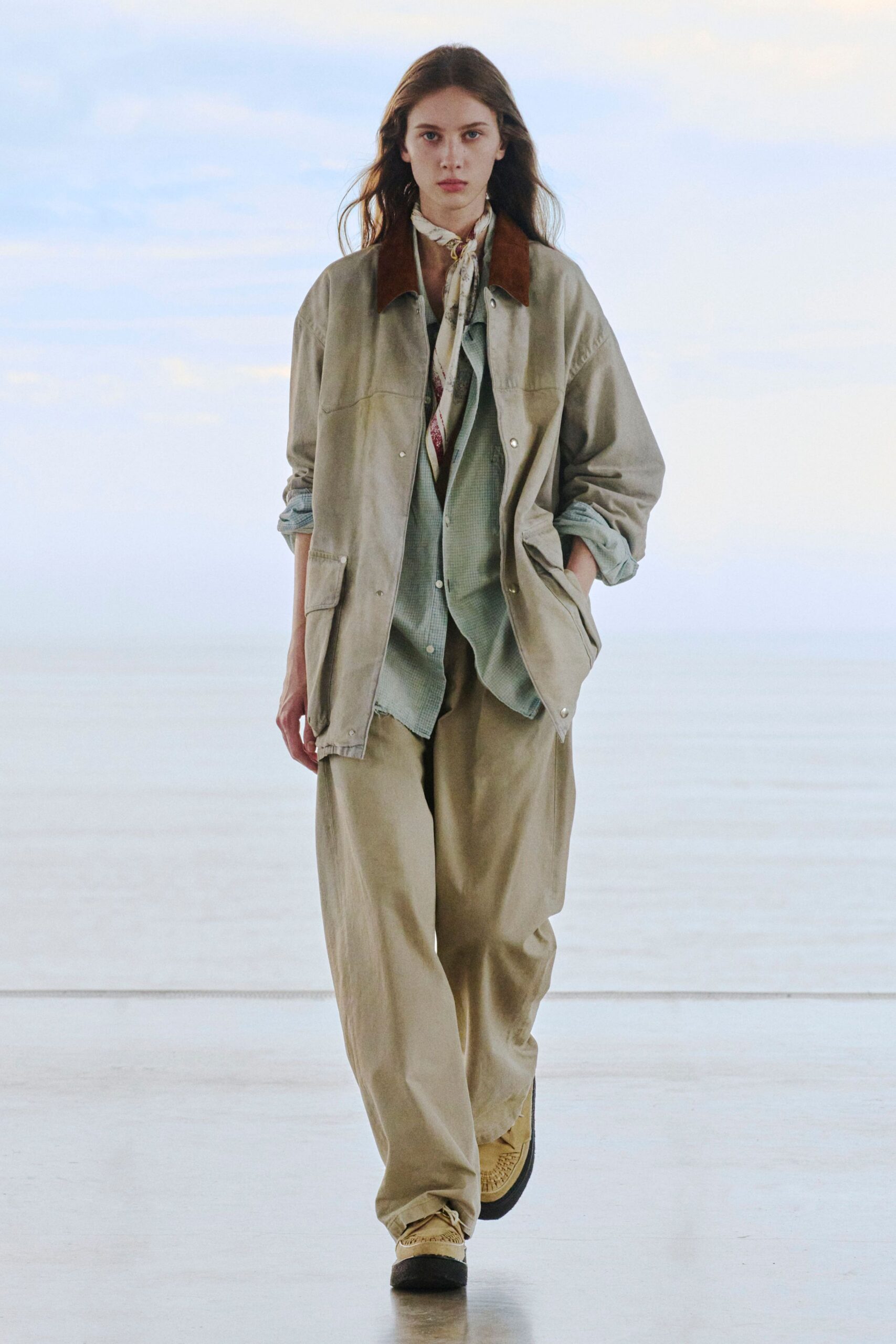 Stone field jacket with a dark suede collar and snap placket, over a pale sage micro-check shirt left loose at the hem. Cream silk scarf knotted close at the throat, sand wide-legs, tan moccasins. Ferry deck at first light.