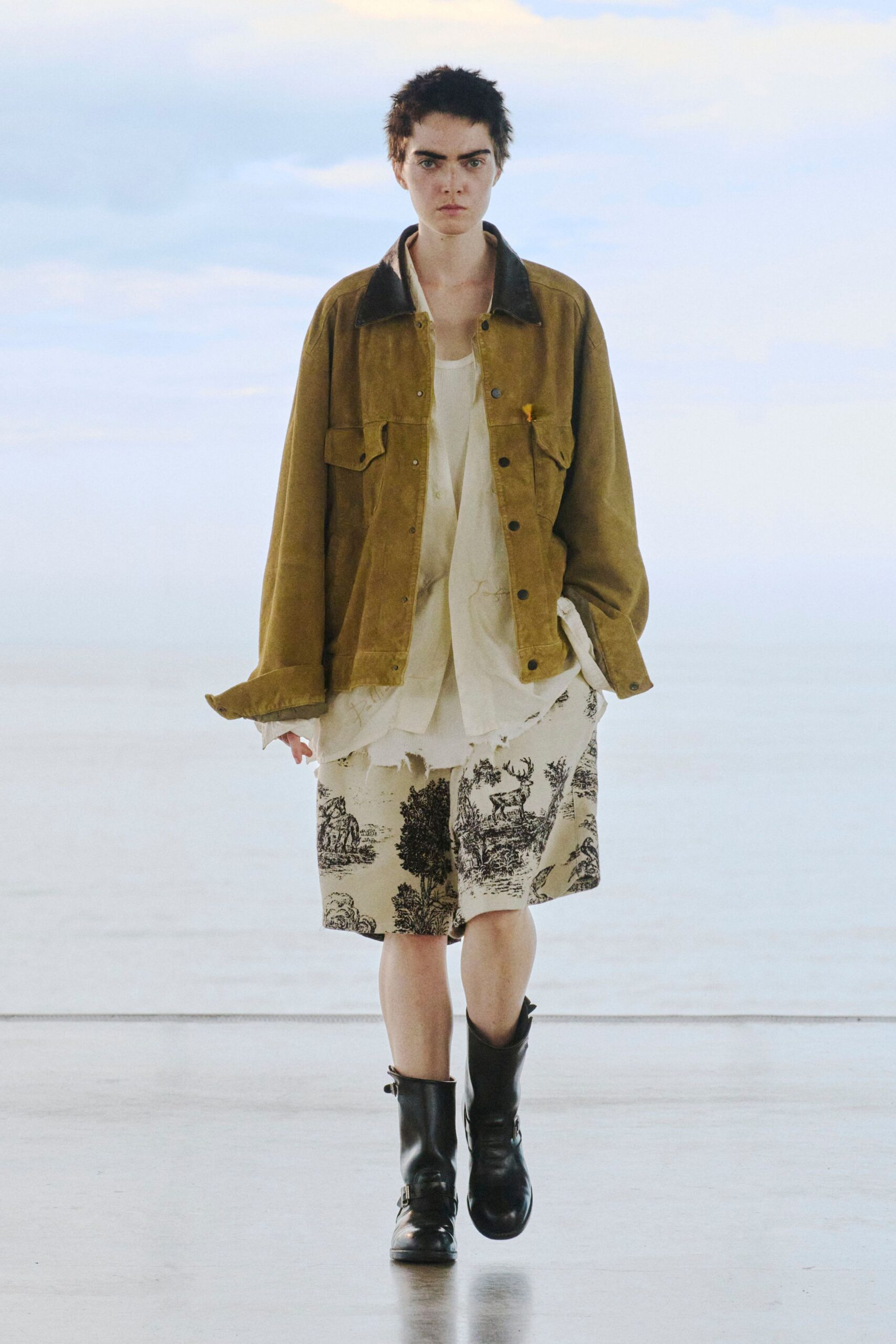 Oversized ochre waxed-cotton field jacket with a dark corduroy collar, over a white ribbed tank and frayed white shirt. Cream knee-length shorts printed like toile, black harness boots. Cold morning at a roadside inn.